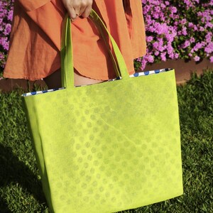 May include: A bright green tote bag with a white polka dot pattern. The bag has a blue and white checkered lining and is being held by a person wearing an orange shirt.