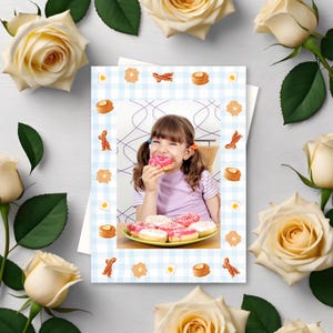 May include: A greeting card with a photo of a young girl eating a pink donut, surrounded by a blue and white checkered border with food illustrations. The card is framed by cream-colored roses.