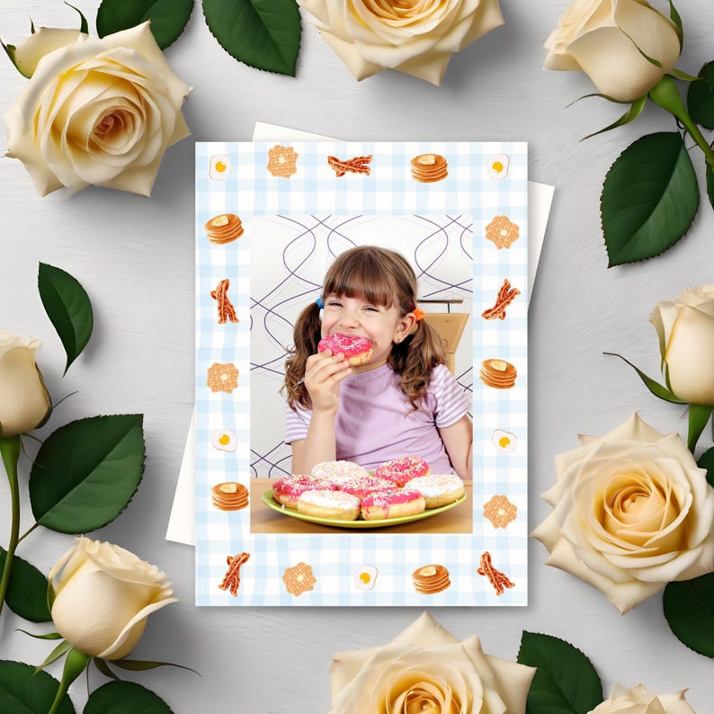 May include: A greeting card with a photo of a young girl eating a pink donut, surrounded by a blue and white checkered border with food illustrations. The card is framed by cream-colored roses.