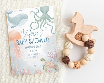 May include: A baby shower invitation with watercolor illustrations of marine life, including a whale, octopus, jellyfish, and sea turtle. The invitation reads "Valerie's BABY SHOWER" with the date and time. A wooden rocking horse teether and a beaded ring are also in the image.