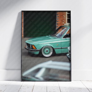 May include: A framed print featuring a vintage teal car. The car has a distinctive design with a chrome bumper and custom wheels. The background includes a green garage door and brick wall, creating a classic automotive aesthetic.