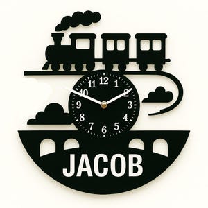 Puede incluir: Reloj de pared negro con temática de tren con el nombre "JACOB" en la parte inferior. La esfera del reloj es negra con números y manecillas blancas. El tren y el puente también son negros, creando un diseño contrastante sobre un fondo blanco.
