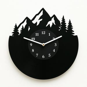 Può includere: Orologio in vinile nero con un design a silhouette di montagna e albero. Il quadrante dell'orologio ha numeri e lancette bianchi.