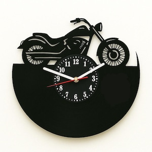 Puede incluir: Un reloj negro con silueta de motocicleta y manecillas blancas. La esfera del reloj presenta números blancos y un segundero rojo. El diseño de la motocicleta está montado sobre una base negra semicircular.