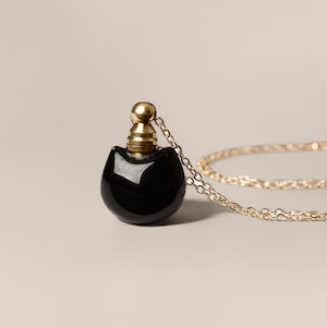 Puede incluir: Un colgante negro con forma de botella de perfume de gato con tapón y cadena dorados. El colgante está hecho de un material oscuro y brillante y cuelga de una delicada cadena dorada. El diseño es minimalista y elegante.