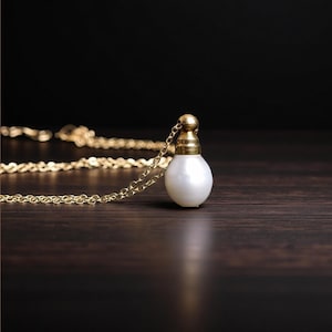 Pode incluir: Um colar de corrente dourada com um pequeno pingente branco perolado em forma de bulbo. O pingente tem uma tampa dourada. O colar é exibido em uma superfície de madeira marrom escura.