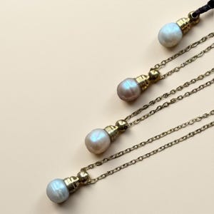 May include: Four pearl pendant necklaces with gold-colored chains and clasps. Each necklace features a lustrous, iridescent pearl suspended from a decorative gold-toned metal piece. The necklaces are arranged diagonally against a light beige background.