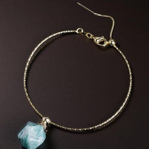 May include: A gold-colored necklace with a textured band and a clasp. The necklace features a pendant with a faceted, blue-green gemstone. The necklace is displayed on a dark brown surface.