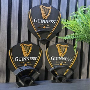 May include: Three black Guinness dart holders with the Guinness logo, a gold harp, and text. The holders are mounted on a wall with a vertical wood panel design. The holders have a black and grey design.