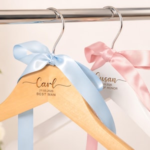 May include: Two wooden clothes hangers with silver metal hooks. One hanger is natural wood with a light blue satin ribbon and engraved text reading Carl, 21.05.2026, BEST MAN. The other is white with a pink satin ribbon and engraved text reading Susan, 21.05.2026, MAID OF HONOR.