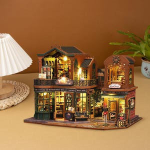May include: A miniature wooden dollhouse model of a two-storey building with a green roof and brown brick facade. The building has two shop fronts, one labelled "Book Shop" and the other "Magic Warcraft". The dollhouse is lit with small LED lights and has a detailed interior.
