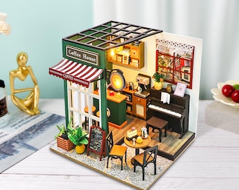 Peut inclure: Une maison de poupée miniature représentant un café avec une façade verte, une marquise rayée rouge et blanche et l'inscription "Coffee House" sur la façade. L'intérieur comprend un comptoir, des tables, des chaises, un piano et une fenêtre donnant sur une rue de la ville.