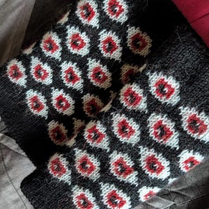 Pode incluir: Polainas de malha pretas com um padrão repetido de formas vermelhas e brancas semelhantes a olhos. As polainas estão dobradas e repousam sobre uma superfície cinza estampada, com um tecido vermelho no fundo.