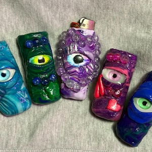 May include: A collection of five uniquely designed lighters, each featuring a sculpted eye motif. The lighters are in various colors, including blue, green, and purple, with intricate details and textures. The eye designs vary in color and style.