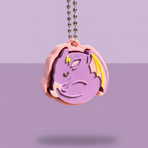 Könnte beinhalten: Ein lila Anhänger mit einem Cartoon-Drachendesign, mit gelben und weißen Akzenten. Der Anhänger ist an einer goldfarbenen Kette befestigt. Der Drache ist vor einem rosa Hintergrund platziert.