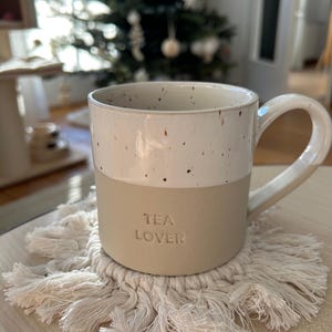 Könnte beinhalten: Eine weiße Keramiktasse mit braunen Flecken und dem Schriftzug "TEA LOVER" an der Seite, die auf einem weißen gehäkelten Untersetzer mit Fransen steht.