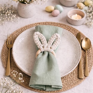 Peut inclure: Mise en place de Pâques avec une serviette vert clair ornée d'un anneau de serviette en forme d'oreilles de lapin. Comprend une assiette blanche, des couverts dorés et un dessous de plat tressé. Bougies et fleurs.