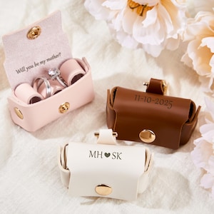 May include: Three small leather ring boxes in pink, brown, and white. The pink box is open, revealing rings and the text "Will you be my mother?" The brown box has "11-10-2025" and the white box has "MH ❤️ SK" printed on them. Gold hardware.