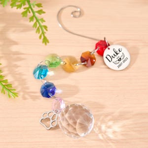 May include: A memorial suncatcher with a rainbow of faceted beads and a clear crystal ball. A silver paw print charm and a white tag with the name "Duke" and dates 2007-2023 are attached. The suncatcher hangs from a silver hook.