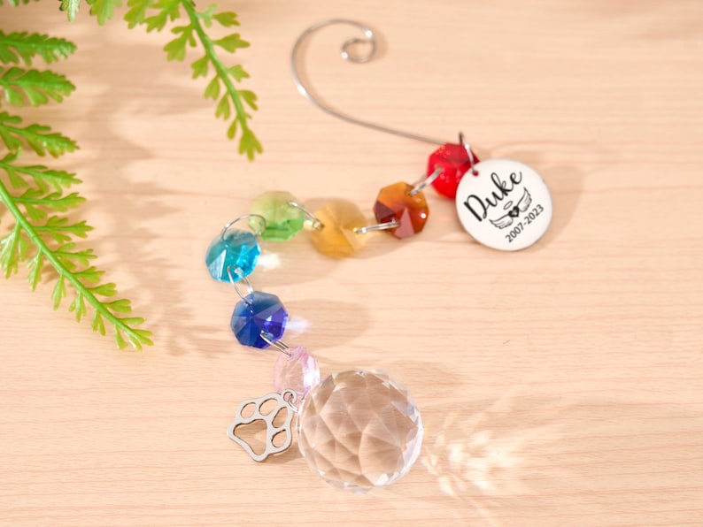 May include: A memorial suncatcher with a rainbow of faceted beads and a clear crystal ball. A silver paw print charm and a white tag with the name "Duke" and dates 2007-2023 are attached. The suncatcher hangs from a silver hook.