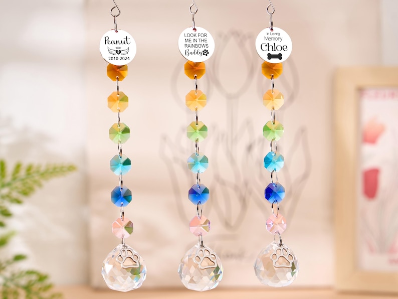 May include: Three memorial suncatchers with rainbow-colored crystal beads and a clear crystal ball with a paw print. Each suncatcher has a circular charm with a pet's name and dates or a message like "Look for me in the rainbows."