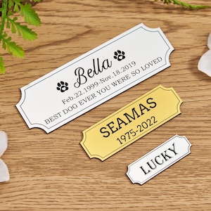 Peut inclure: Trois étiquettes commémoratives personnalisées pour animaux de compagnie. La plus grande est blanche avec du texte noir et des empreintes de pattes, avec le nom "Bella" et des dates. Une étiquette dorée indique "Seamas" et des dates. Une petite étiquette blanche indique "Lucky".