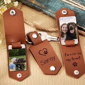Puede incluir: Llaveros de cuero marrón con inserciones de fotos metálicas. Un llavero tiene el nombre "Saray" con un corazón y una huella de pata. Otro dice "Forever in my heart" con una huella de pata. Los llaveros presentan fotos de una niña y un perro.
