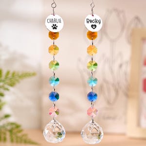 May include: Two hanging crystal suncatchers with the names "Charlie" and "Rocky" on round silver charms. Each suncatcher features a paw print, a series of colorful crystals, and a clear crystal teardrop with a paw print.