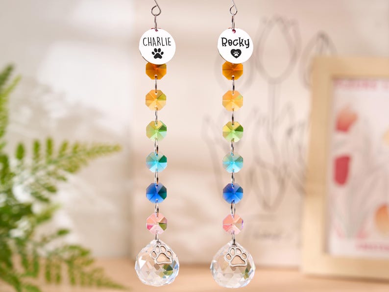 May include: Two hanging crystal suncatchers with the names "Charlie" and "Rocky" on round silver charms. Each suncatcher features a paw print, a series of colorful crystals, and a clear crystal teardrop with a paw print.