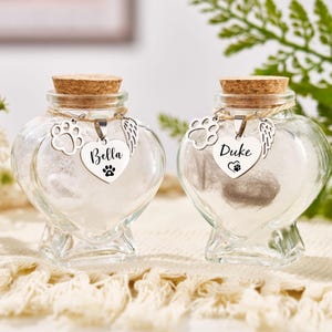 Op de afbeelding: Twee hartvormige glazen urnen met kurken stoppers, elk met een pootafdrukbedel en een naamlabel met de tekst "Bella" en "Duke". De urnen zijn gevuld met een witte substantie, waarschijnlijk as.