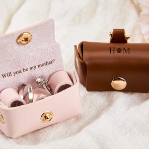 May include: Two leather ring boxes. One is pink and open, displaying rings and the text "Will you be my mother?" The other is brown, closed, with the initials "H & M" and a paw print. Both have gold hardware.