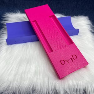 May include: A bright pink rectangular tool with the text D3-3D embossed on the surface, featuring a central recessed channel with measurement markings, positioned over a curved purple base component with visible numerical scale markings.