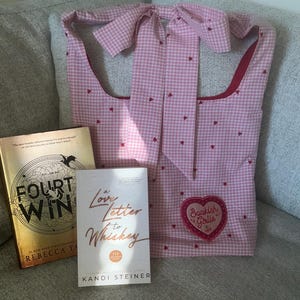 May include: A pink gingham tote bag with red heart accents and a heart-shaped patch that says "Bookish Girlie." Two books are next to the bag: "Four Win" and "A Love Letter to Whiskey."