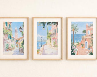 May include: A set of three framed watercolour prints showcasing Italian coastal scenes. Each print presents a unique view: a narrow street with a church tower, a coastal road with a mountain backdrop, and a seaside villa with palm trees. The frames are light-coloured wood.