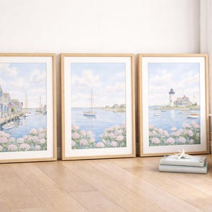 May include: Three framed watercolor prints depicting coastal scenes. The artwork features a harbor with boats, a sailboat on the water, and a lighthouse. The prints are in light wood frames, resting on a wooden floor.