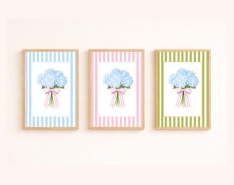 May include: Three framed floral prints showcasing blue hydrangea bouquets with pink bows. Each print has a different striped border: blue, pink, and green. The frames are made of light-coloured wood, creating a coordinated set.