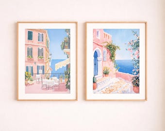 May include: Two framed watercolour prints showcase Italian coastal scenes. One depicts a pink building with a balcony and a dining table. The other features a pink building with a blue door and a sea view. Both are framed in light wood.