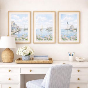 May include: Three framed watercolor prints depicting coastal scenes with boats, buildings, and a lighthouse. The prints are displayed above a white desk with a woven lamp, flowers, and a striped chair. The room has white shelves and woven baskets.