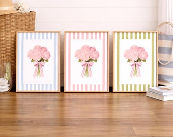 May include: Three framed floral prints showcasing pink hydrangea bouquets tied with pink ribbons. Each print has a striped border in blue, pink, and green. The frames are light wood, displayed on a wooden floor.