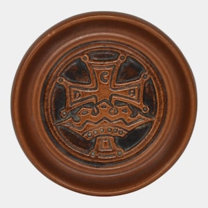 May include: A round, brown ceramic dish with a raised rim. The center features a detailed, embossed design of a cross, crown, and decorative elements. The dish has a rustic, antique appearance.