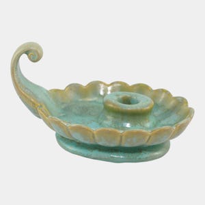 May include: A ceramic candle holder in a teal and tan color scheme. The holder has a scalloped edge and a curved handle. The candle holder is approximately 6 inches long and 3 inches wide.