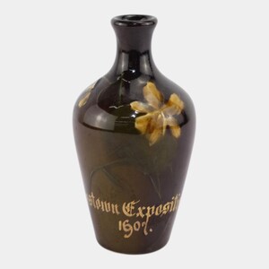 May include: A dark brown ceramic vase with a narrow neck and a rounded body. The vase features a floral design in shades of yellow and brown. The words "town Expositi 1907" are written in gold script on the vase.