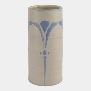 May include: A cylindrical ceramic vase in a light beige color with a blue floral design. The vase has a smooth surface and a slightly flared rim. The blue design is symmetrical and runs vertically along the vase.