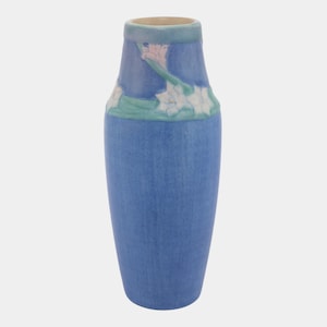 May include: A blue ceramic vase with a floral design. The vase has a cylindrical shape with a slightly flared top. The floral design features white flowers with green and pink accents. The vase is approximately 8 inches tall.