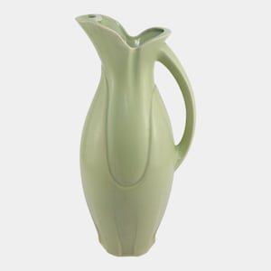 May include: A light green ceramic pitcher with a curved handle and a unique, petal-like spout. The pitcher has a smooth, glossy finish and a rounded body with vertical, subtly raised lines. The overall design is elegant and minimalist.