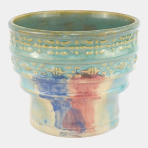May include: A handcrafted ceramic vase with a wide, cylindrical shape. The vase features a turquoise glaze with embossed gold detailing around the top. The lower portion has a blend of blue, red, and cream-colored paint. A unique piece for home decor.