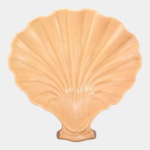 May include: A peach-colored, ceramic seashell-shaped serving dish. The dish has a detailed, ribbed texture radiating from the center, mimicking the natural form of a scallop shell. The base of the shell has a small, rounded stem.