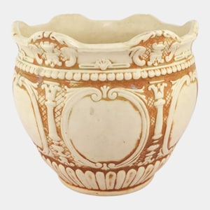 May include: Cream-colored ceramic planter with ornate, raised relief detailing. The planter features a scalloped rim and a repeating pattern of decorative elements in a warm brown hue. The design includes cartouches, columns, and floral motifs.