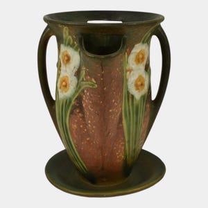May include: A brown and green ceramic vase with two handles and a circular base. The vase features a floral design of white and yellow daffodils with green stems and leaves. The vase has a unique shape with a cutout near the top.