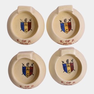 May include: Four round, off-white ceramic ashtrays featuring a central crest with a skull and crossbones, a helmet, and the letters F, C, and B. Each ashtray is printed with the red text K. OF P. GRAND LODGE, with some including ZANESVILLE, OHIO.